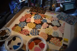 Philip Jacobs design bord, hvor han maler alle sine stofdesigns, inden de bliver trykt til stoffer som i baggrunden. 3 plastik tallerkner, med blandede farver til at male designet med blomster